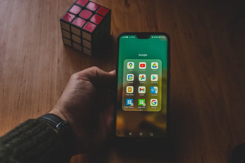 Hand holding an Android phone showing a folder of apps, with a Rubik’s cube on a wooden desk