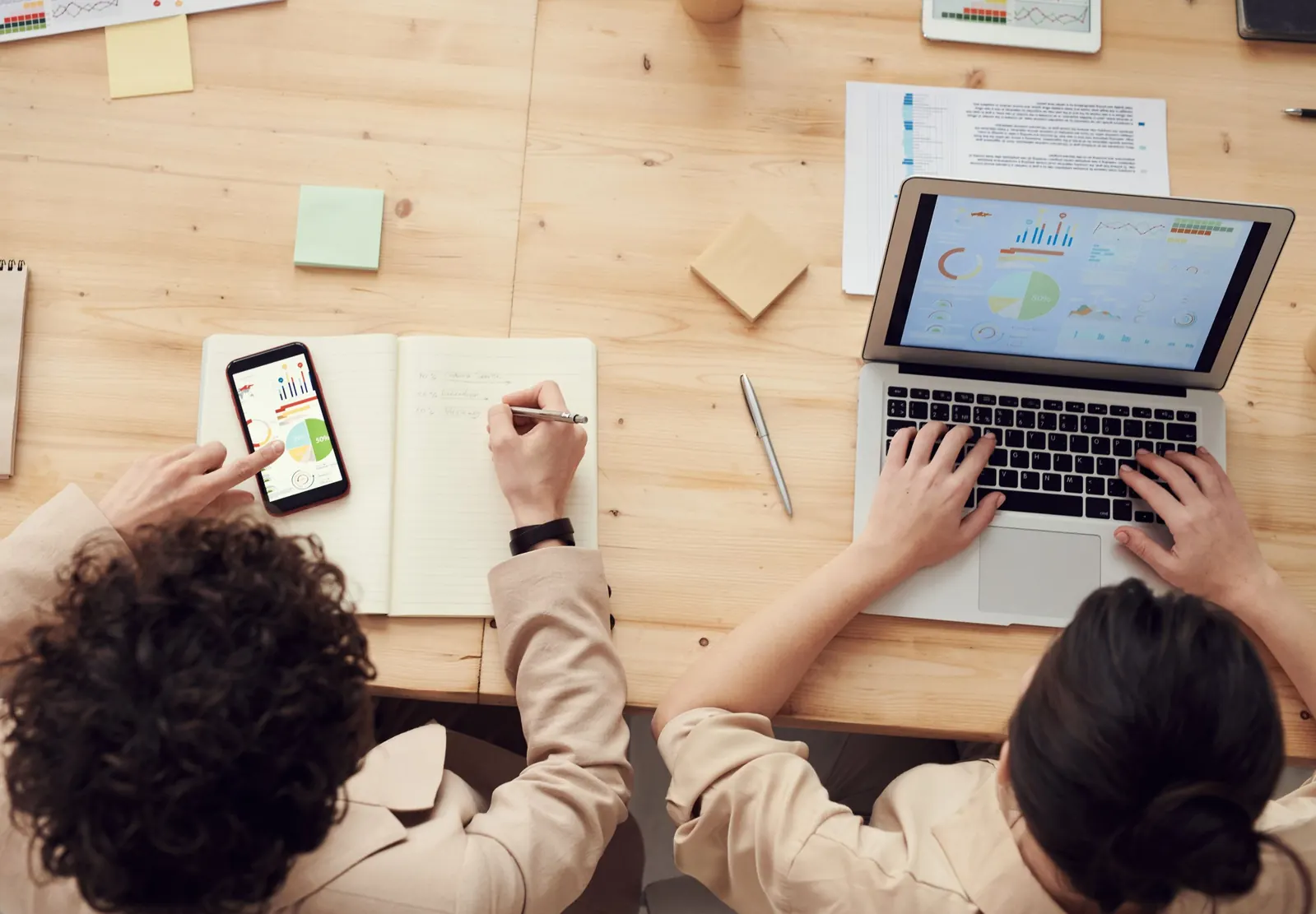 Two people at a wooden desk reviewing analytics dashboards on a laptop and a phone